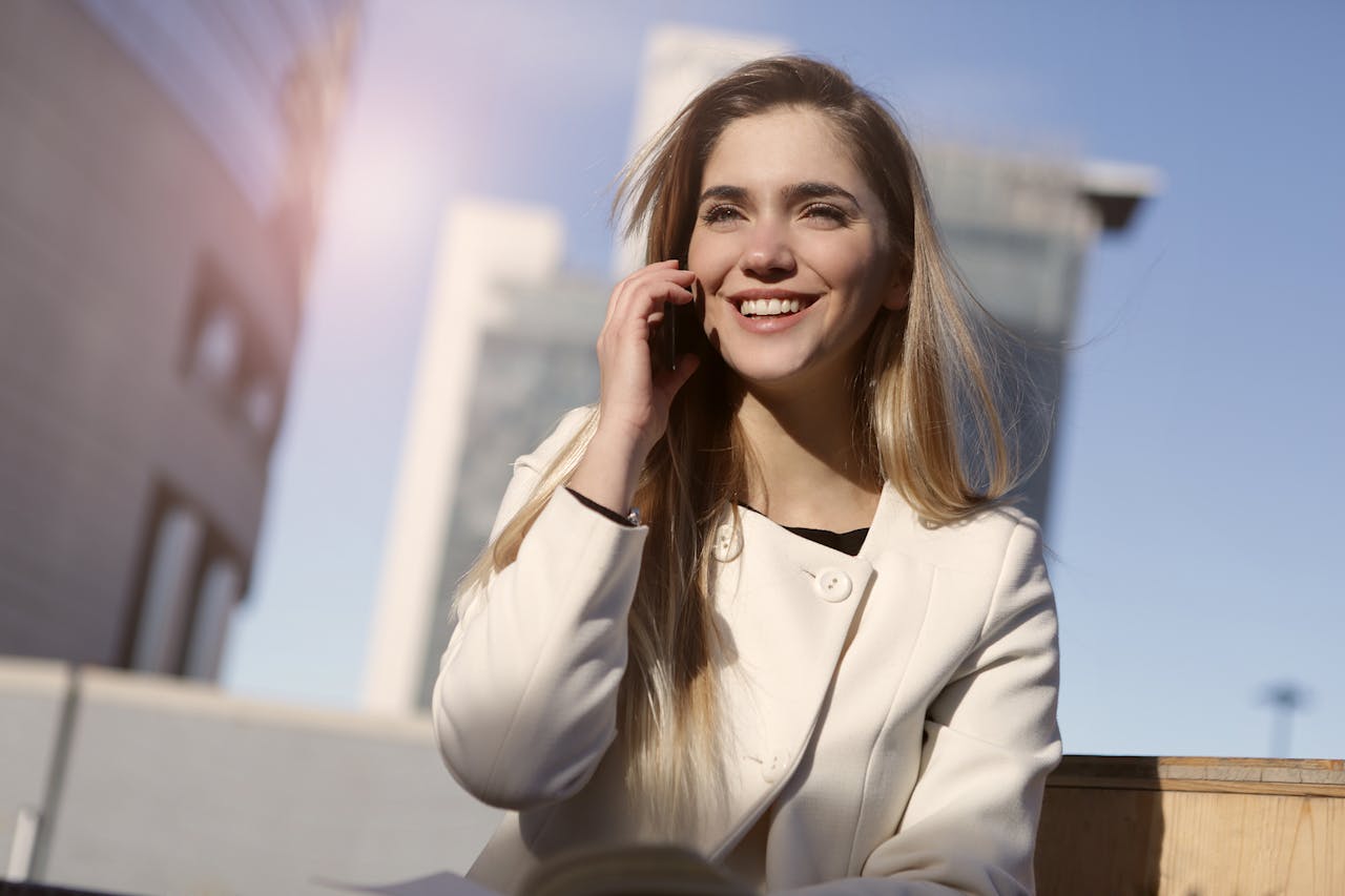 Confident woman in white coat smiling while using a smartphone outside on a sunny day.